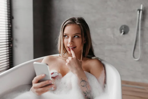 Misterioasă femeie caucaziană care folosește telefonul mobil în timp ce stă întinsă în baie spumoasă, trimite mesaje text online, gândește și privește deoparte — Fotografie, imagine de stoc
