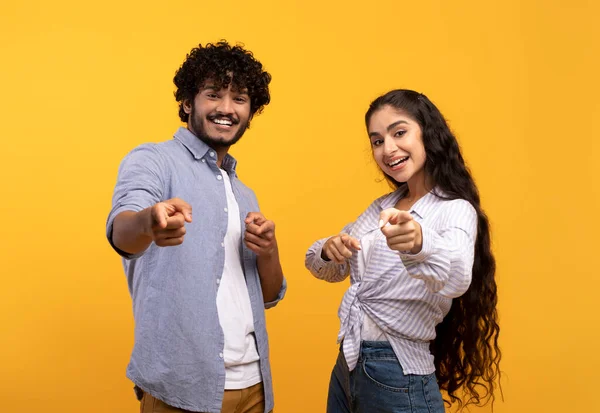 ¿Y qué hay de ti? Emocionada pareja india apuntando con los dedos índice a la cámara, posando sobre fondo amarillo del estudio — Foto de Stock