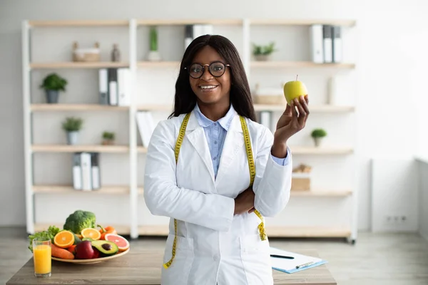 Alegre consejera de nutrición afroamericana con manzana y cinta métrica trabajando en clínica y sonriendo a la cámara —  Fotos de Stock