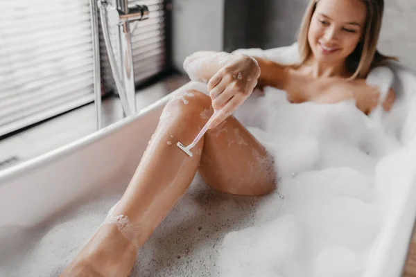 Jovencita acostada en un baño espumoso y afeitándose las piernas con maquinilla de afeitar, disfrutando de un procedimiento de spa doméstico, haciendo depilación —  Fotos de Stock