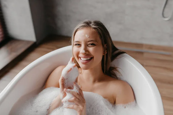 Relaksująca kąpiel. Szczęśliwa pani leży w wannie, korzystając z rytuału piękna, patrząc i uśmiechając się do kamery, przestrzeń kopiowania — Zdjęcie stockowe