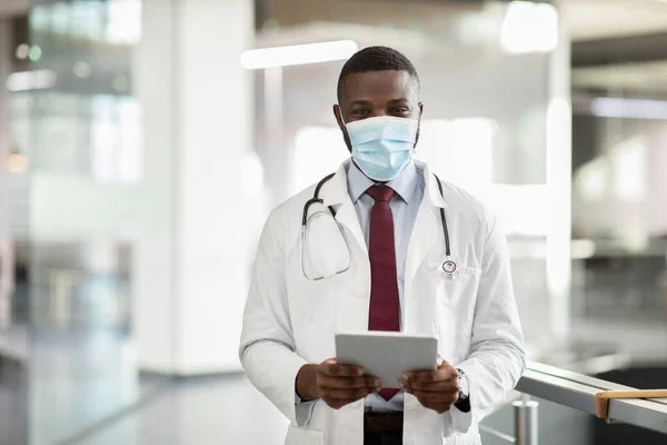 Médico preto na máscara facial segurando tablet digital, clínica interior — Fotografia de Stock