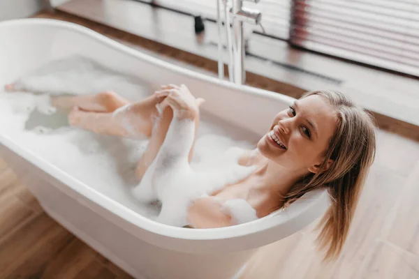 Mujer joven y relajada tomando un baño con espuma, sonriendo a la cámara, descansando en la bañera de hidromasaje en la mañana en casa —  Fotos de Stock