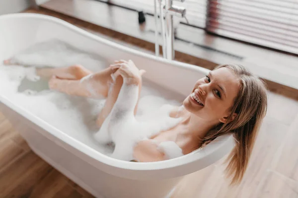 Jeune femme détendue prenant un bain avec de la mousse, souriant à la caméra, se reposant dans un bain à remous le matin à la maison — Photo