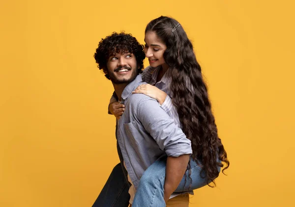 Güzel Hintli kadın erkek arkadaşını sırtında taşıyor, gülümseyen adam kız arkadaşını sırtında taşıyor, sarı arka plan. — Stok fotoğraf