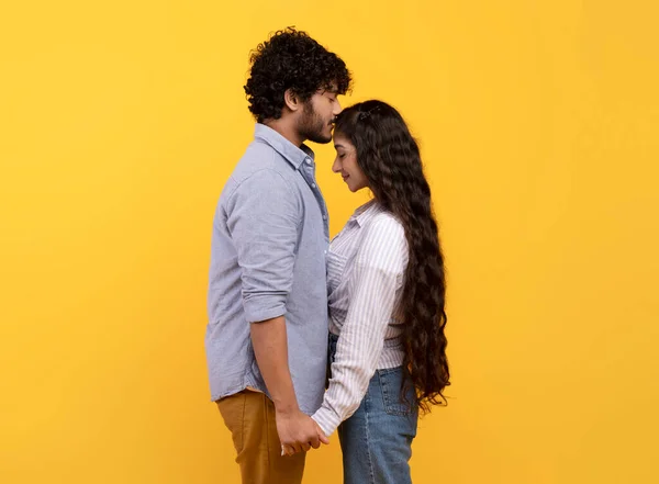 Ніжний індійський чоловік дарує своїй дівчині поцілунок на лобі, висловлюючи свою любов, стоячи над жовтим тлом — стокове фото