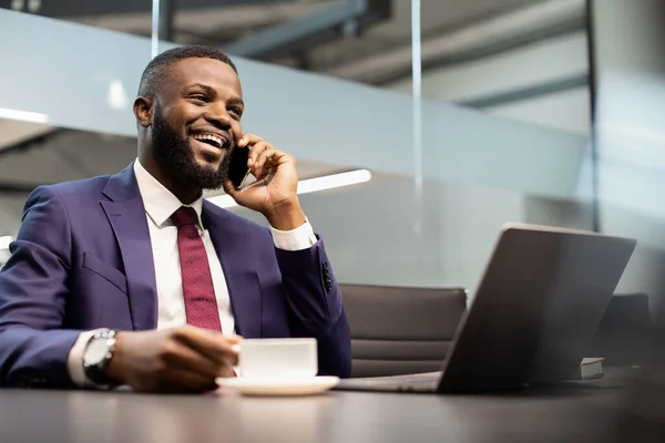 Negocio afroamericano positivo que trabaja en el ordenador portátil, teniendo conversación telefónica — Foto de Stock