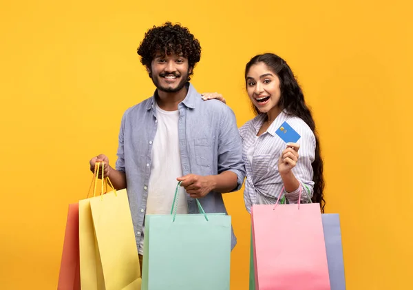 Concepto de consumismo. Emocionada pareja india con tarjeta de crédito y bolsas de compras, compra remota, fondo amarillo — Foto de Stock