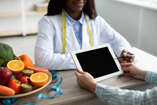 검은 여성 영양학자는 빈 화면으로 태블릿을 가리키면서, 식사 계획을 설명하고 환자에게 조언을 베 푼다 — 스톡 사진
