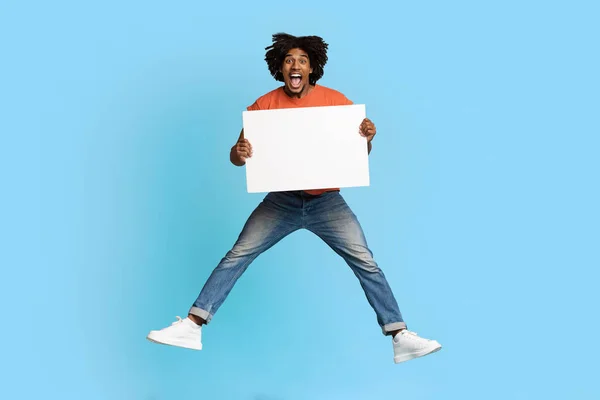 Емоційний чорний хлопець стрибає з порожнім плакатом над синім — стокове фото