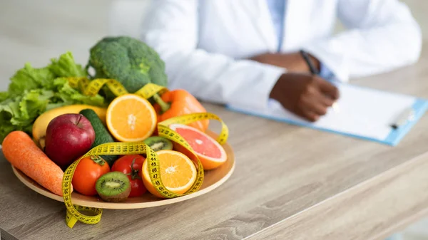 Чорний дієтолог пише план дієти, сидячи на робочому місці в клініці, зосереджуючись на тарілці з фруктами та овочами, обрізаними — стокове фото