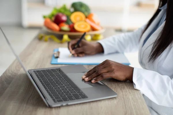 Рабочий процесс. Африканский американский диетолог разрабатывает план диеты для пациента или подсчета калорий, работает на ноутбуке — стоковое фото