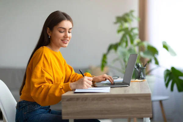 Mujer caucásica feliz usando PC portátil, trabajando o estudiando en línea, tomando notas durante el webinar o lección remota en casa — Foto de Stock