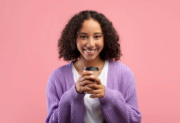 Glad ung svart kvinna som håller takeaway kaffe i engångs pappersmugg och ler mot kameran på rosa bakgrund — Stockfoto