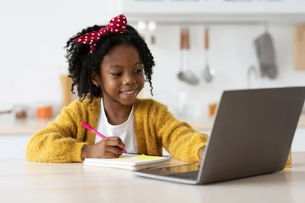 Bonito preteen preto menina escrita no bloco de notas enquanto estudo on-line com laptop — Fotografia de Stock