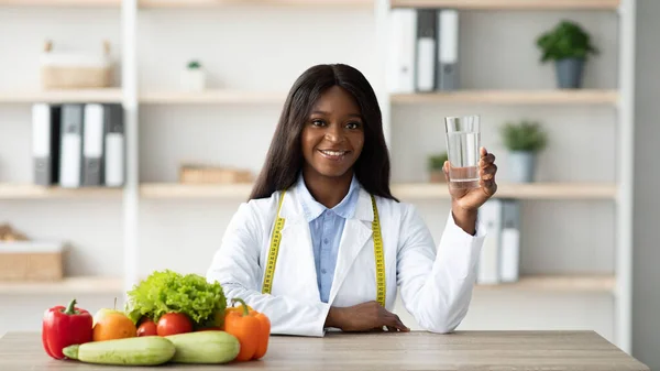 Jeune nutritionniste afro-américaine tenant un verre d'eau assis à table avec des fruits et légumes — Photo