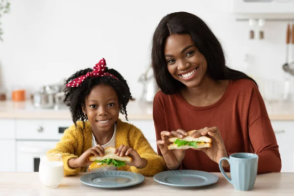 Retrato de feliz negro madre y poco hija comiendo sándwiches en cocina — Foto de Stock