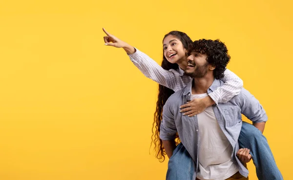 Entusiasmado jovem casal indiano se divertindo e senhora piggybacking cara e apontando o dedo para o espaço livre no fundo amarelo — Fotografia de Stock