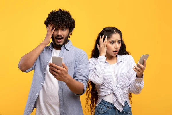 Femme indienne et son petit ami regardant dans leurs smartphones en état de choc et touchant les têtes sur fond jaune — Photo