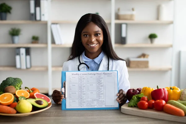 Ernährungskonzept. Junge Diätassistentin zeigt wöchentlichen Ernährungsplan, sitzt am Schreibtisch mit Tellern mit Obst und Gemüse — Stockfoto