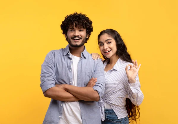 Retrato de feliz pareja india abrazando, mujer mostrando gesto bien y sonriendo a la cámara sobre fondo amarillo — Foto de Stock