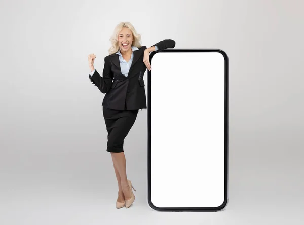 Ja. Überglückliche Geschäftsfrau lehnt an riesigem Smartphone mit leerem Bildschirm und ballt geballte Faust, Attrappe — Stockfoto