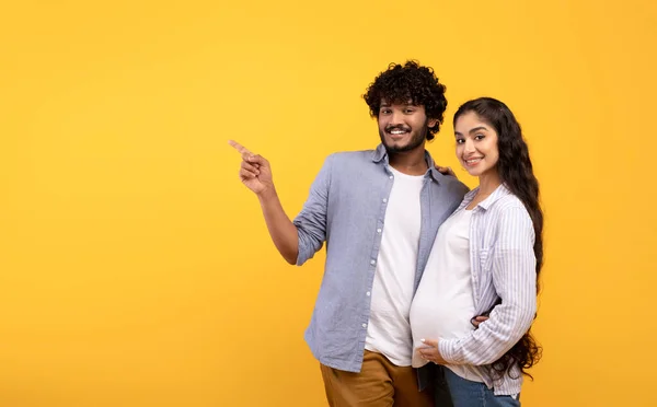 Νέοι αγαπώντας ινδική έγκυος ζευγάρι επισημαίνοντας κατά μέρος το χώρο αντίγραφο, δείχνοντας μεγάλη προσφορά ή promo, κίτρινο φόντο — Φωτογραφία Αρχείου
