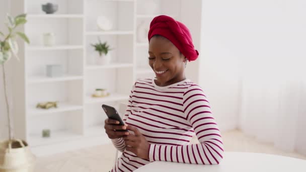 Alegre negro hembra usando Headwrap usando el teléfono en casa — Vídeo de stock