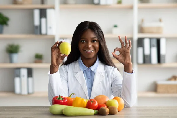 Giovane dietista nero in possesso di mela, mostrando gesto ok, seduto alla scrivania con frutta e verdura, sorridente alla macchina fotografica — Foto Stock