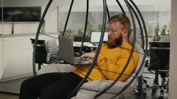 Chico usando computadora portátil escribiendo sentado en silla de huevo en la oficina — Vídeo de stock