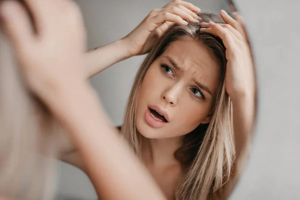 灰心丧气的女人在洗澡间的镜子里看着自己的倒影，寻找有头皮屑问题的发片 — 图库照片