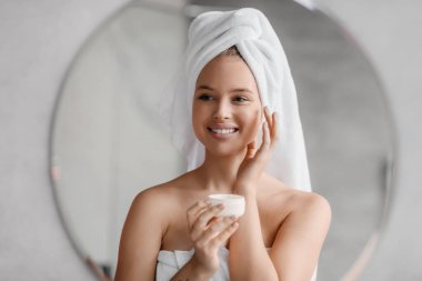 Mutlu genç bayan yüz kremi kullanıyor, aynaya bakıyor ve gülümsüyor, yanaklarına güzellik ürünü sürüyor.