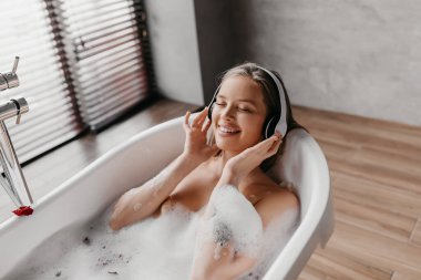Çekici bir kadın sıcak bir banyoda uzanıyor ve kablosuz kulaklıkla müzik dinliyor, spa gününde müzik listesinin keyfini çıkarıyor.