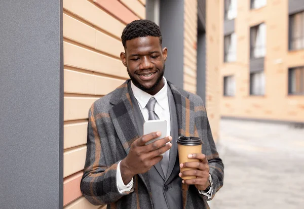 Ευτυχισμένος Αφροαμερικάνος επιχειρηματίας που κρατάει καφέ και χρησιμοποιεί smartphone ενώ περπατάει στο ύπαιθρο — Φωτογραφία Αρχείου