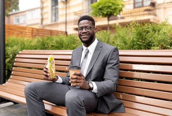 Njut av lunchen. Glad afrikansk affärsman dricker kaffe och äter smörgås, sitter på bänken — Stockfoto