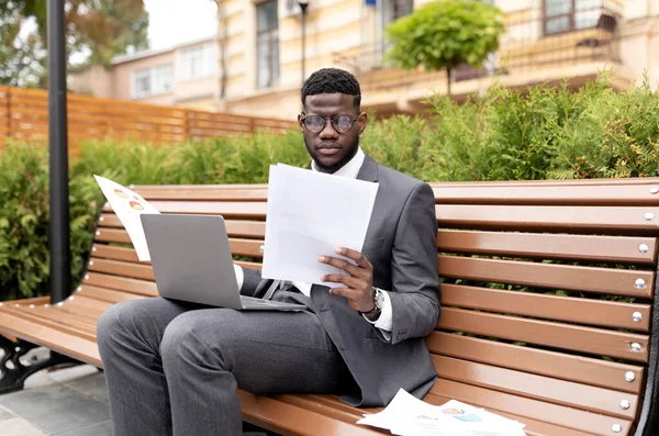 Επιχειρηματική ιδέα καριέρας. Σοβαρός Αφροαμερικάνος επιχειρηματίας που εργάζεται σε laptop και ανάγνωση εγγράφων, κάθεται στον πάγκο — Φωτογραφία Αρχείου