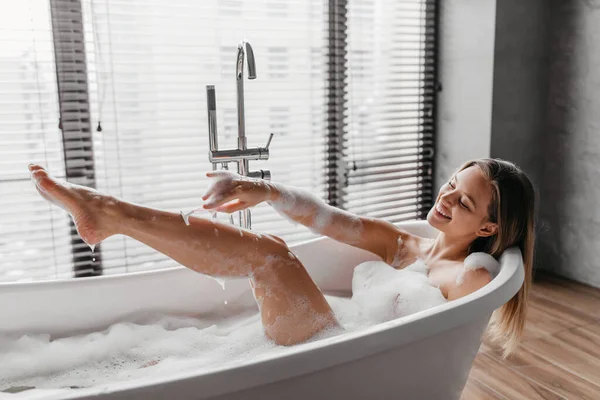 Opieka osobista w domu. Szczęśliwa pani goląca nogi brzytwą, leżąca w piankowej kąpieli z bąbelkami, wykonująca zabieg pielęgnacji skóry — Zdjęcie stockowe