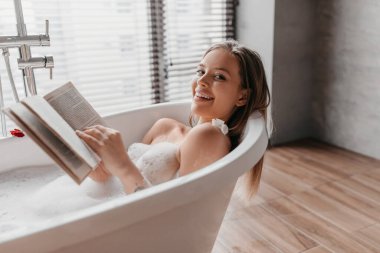 Evdeki banyoda dinleniyor. Köpüklü, kitap okuyan ve kameraya gülümseyen genç bir kadın.