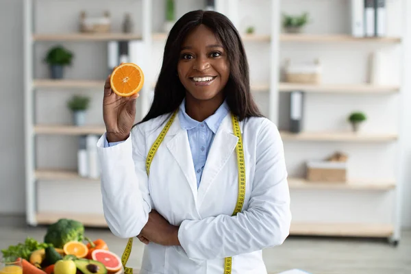 La vitamine C est bonne pour l'immunité. Heureux diétologue afro-américain tenant la moitié orange, recommandant des fruits frais à manger — Photo