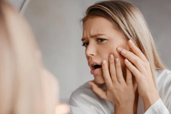 Unhappy lady with popping pimple on cheek while standing near mirror in bathroom at home, young woman suffering acne — Stock Photo, Image