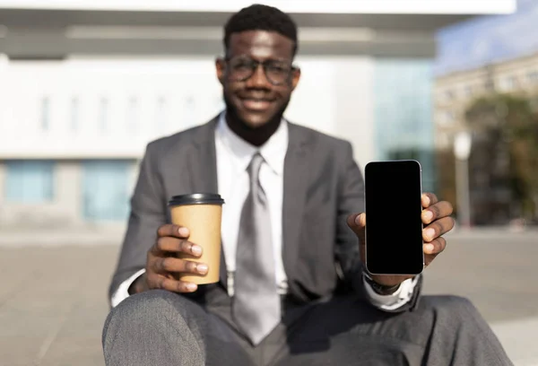 Χαρούμενος Αφροαμερικάνος επιχειρηματίας που επιδεικνύει smartphone με άδεια οθόνη, κάθεται σε εξωτερικούς χώρους με καφέ για να πάει — Φωτογραφία Αρχείου