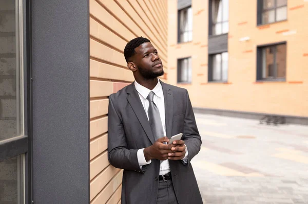 Pensamento estratégico. Pensativo empresário afro-americano segurando smartphone e olhando para longe, apoiando-se na parede — Fotografia de Stock