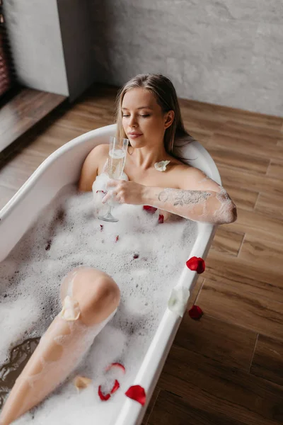 Relaxado jovem bebendo champanhe na banheira de bolhas, comemorando férias com bebida alcoólica em casa, colheita — Fotografia de Stock