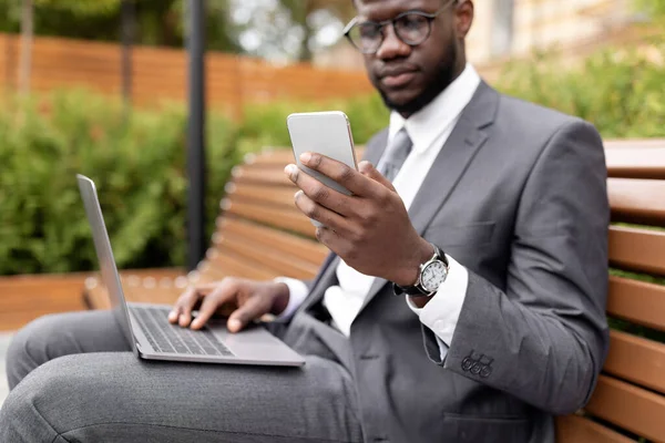 Entrepreneur afro-américain avec ordinateur portable assis sur un banc à l'extérieur et utilisant un smartphone, accent sélectif — Photo