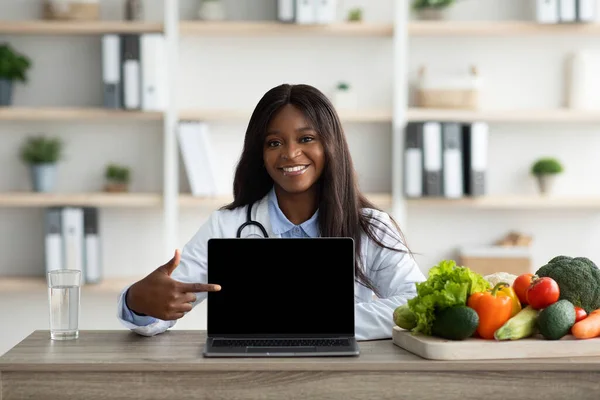Felice specialista di nutrizione africana americana che punta al computer portatile con schermo bianco presso lo studio medico, mockup per il tuo annuncio — Foto Stock
