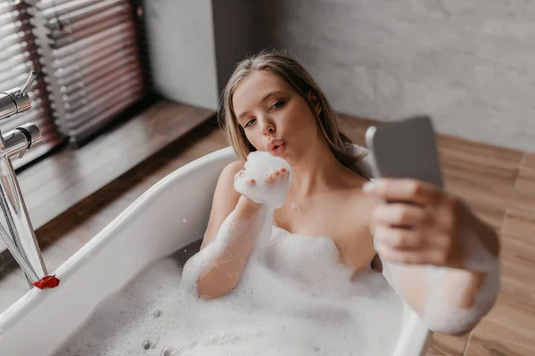 Ładna pani leży w wannie i robi selfie na smartfonie, odpoczywa w łazience i bawi się pianką — Zdjęcie stockowe