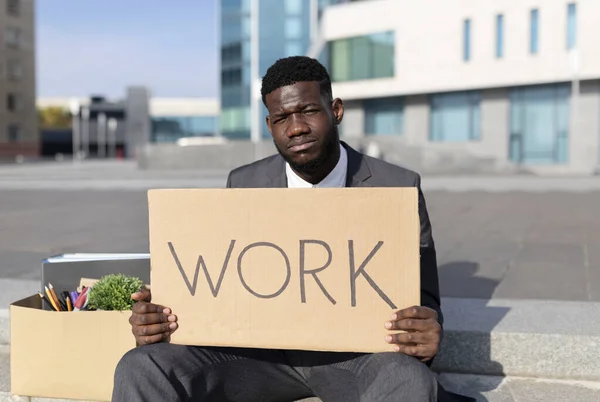 Retrato de empresário negro demitido com cartaz de papel dizendo trabalho e caixa de coisas pessoais, sentado em escadas ao ar livre — Fotografia de Stock