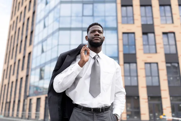 Ung afrikansk amerikansk kontorsarbetare håller jacka på axeln och går nära modern kontorsbyggnad, tittar bort — Stockfoto
