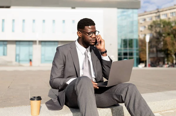 Μαύρος επιχειρηματίας με φορητό υπολογιστή που κάθεται σε εξωτερικούς χώρους και μιλάει σε smartphone, κτίριο γραφείων στο παρασκήνιο, αντιγραφή χώρου — Φωτογραφία Αρχείου
