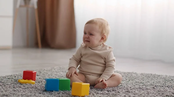 Portrait of cute little baby crying, sitting on floor with colorful cubes, suffering from pain or loneliness, free space — Stock Photo, Image